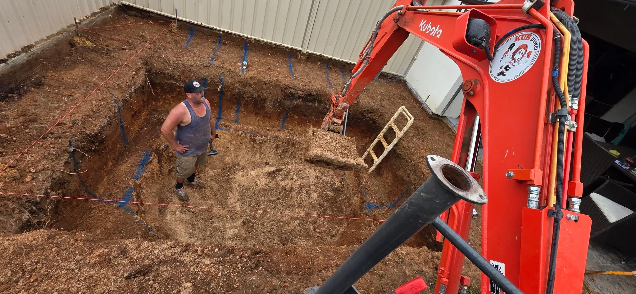 Excavator digging pool hole with string lines and blue marking paint visible at Manningham build site