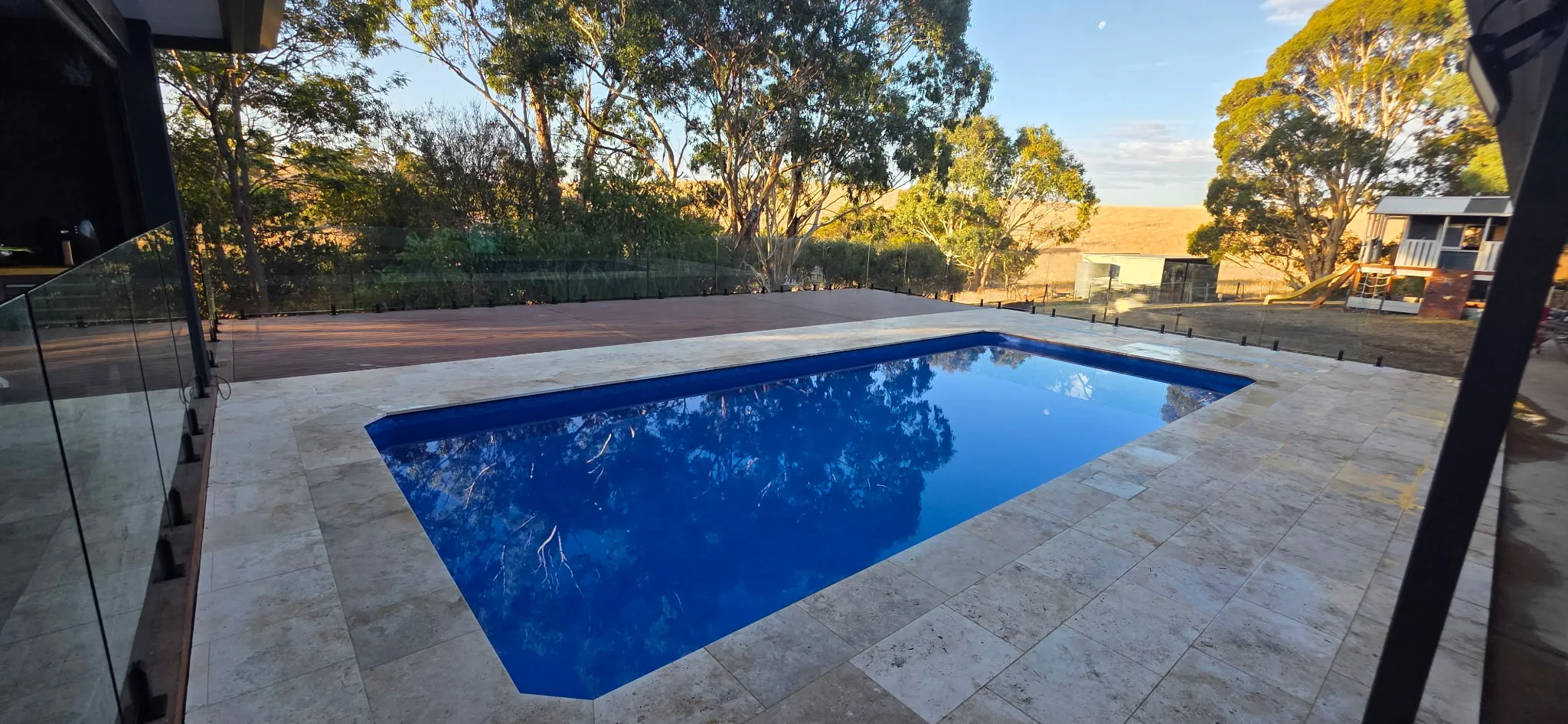 Wide angle daytime view of completed Outback Oasis pool renovation with glass fencing, eucalyptus trees, and golden hillside
