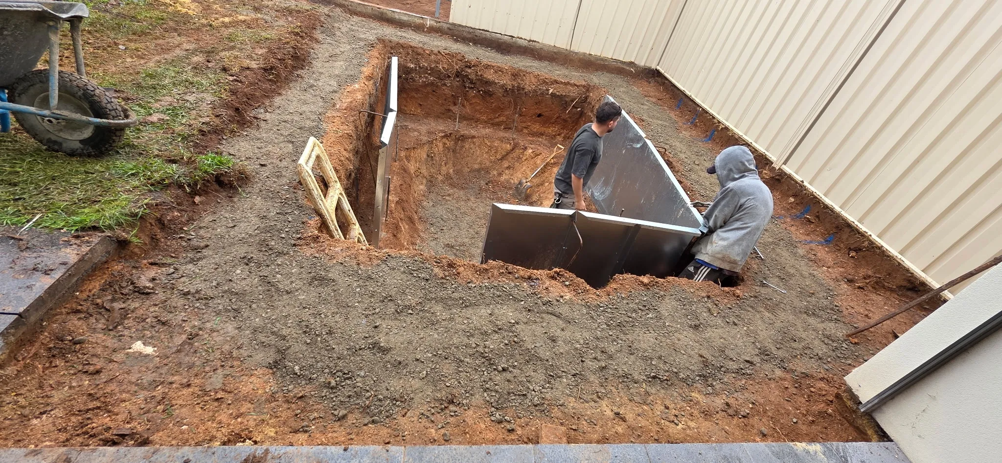 Two workers lowering steel pool wall panels into the excavated hole at Manningham