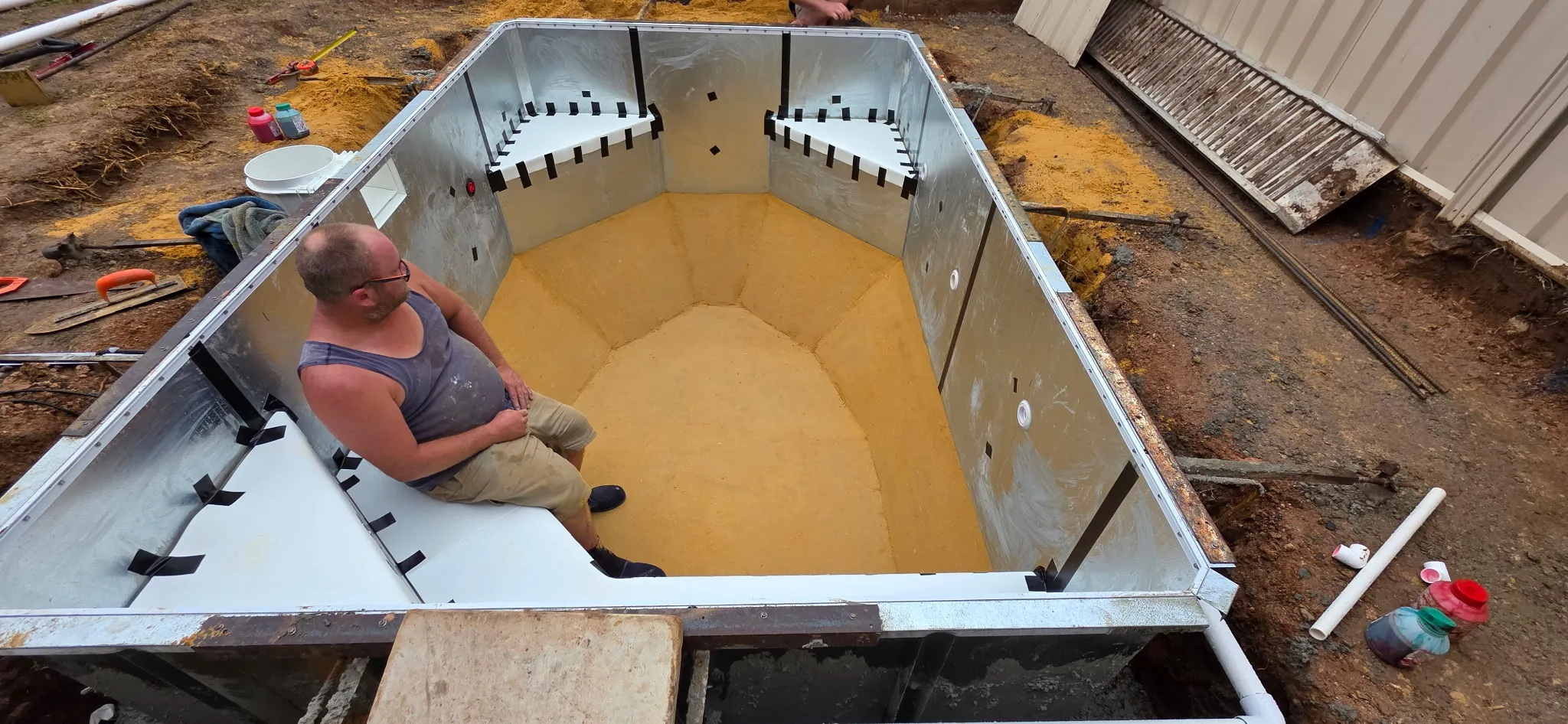 Worker inside steel pool frame smoothing compacted sand base floor before liner installation