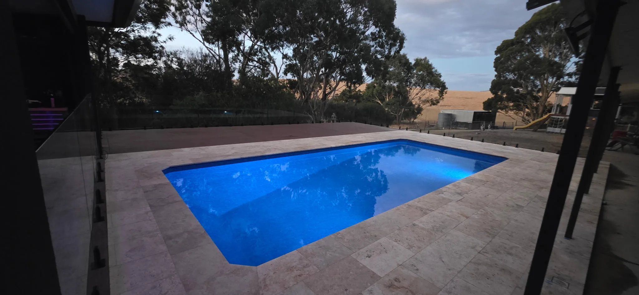 Evening shot of renovated pool with LED lighting illuminating the water through frameless glass fencing with timber deck beyond