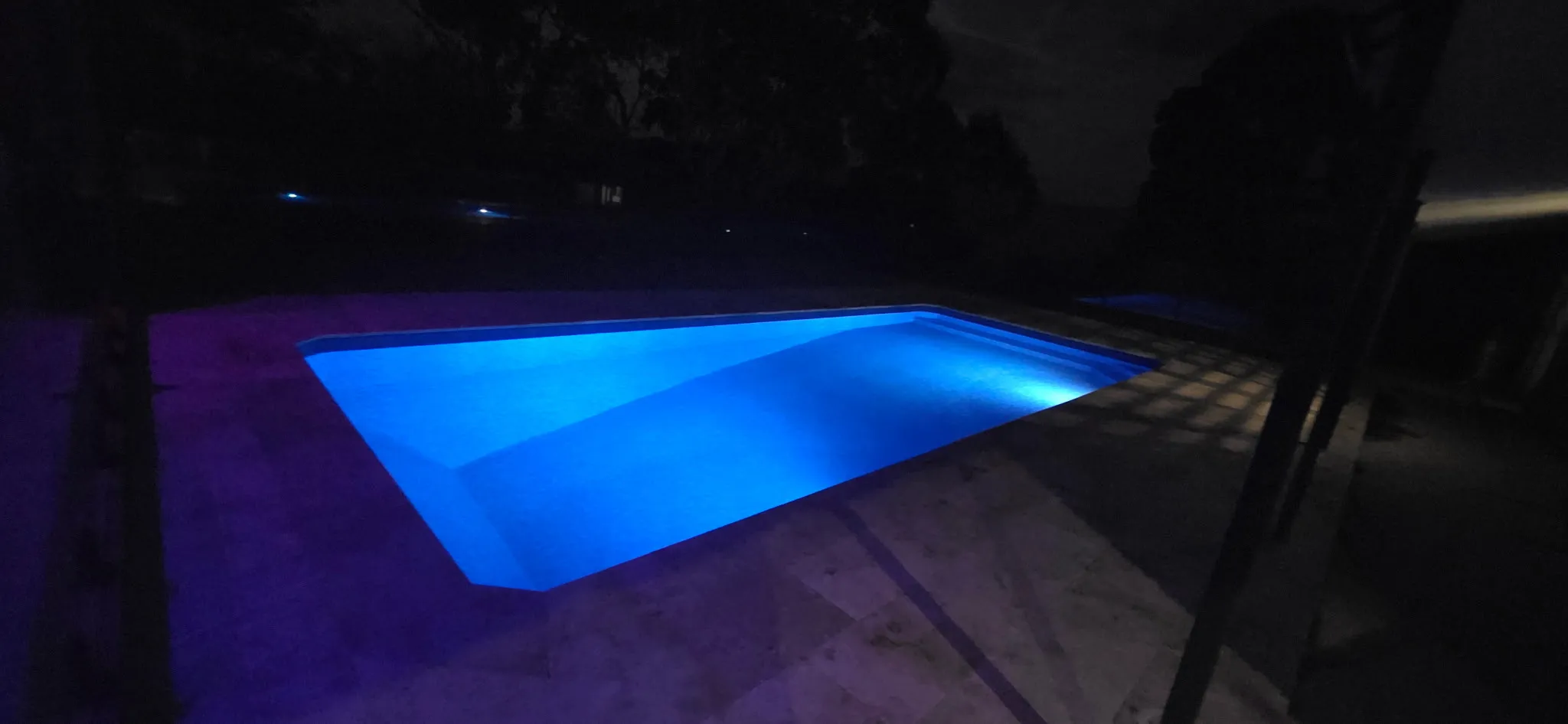 Night shot of Outback Oasis pool with vivid blue LED lighting casting glow across travertine paving and glass fencing