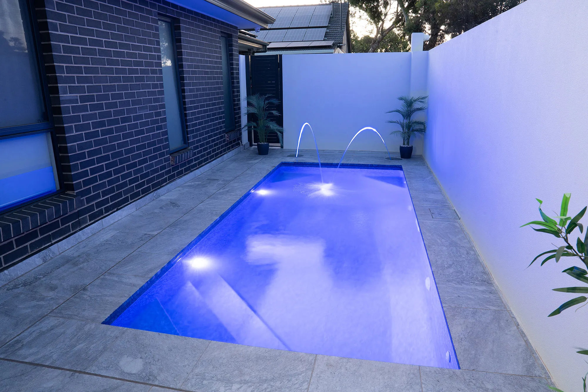 Evening shot of courtyard plunge pool with deep blue LED glow, laminar jets, and reflections on travertine paving