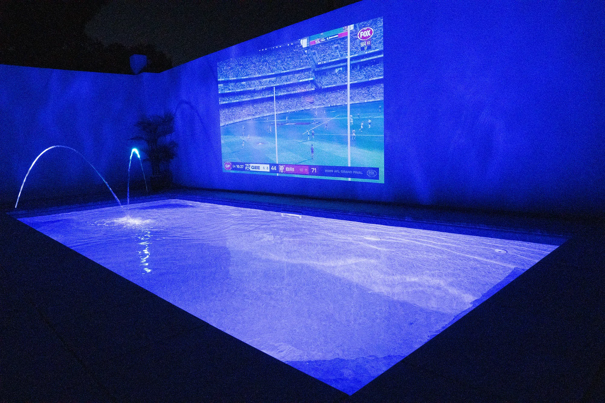 Plunge pool at night with AFL Grand Final projected on rendered white wall above the water with laminar jets running