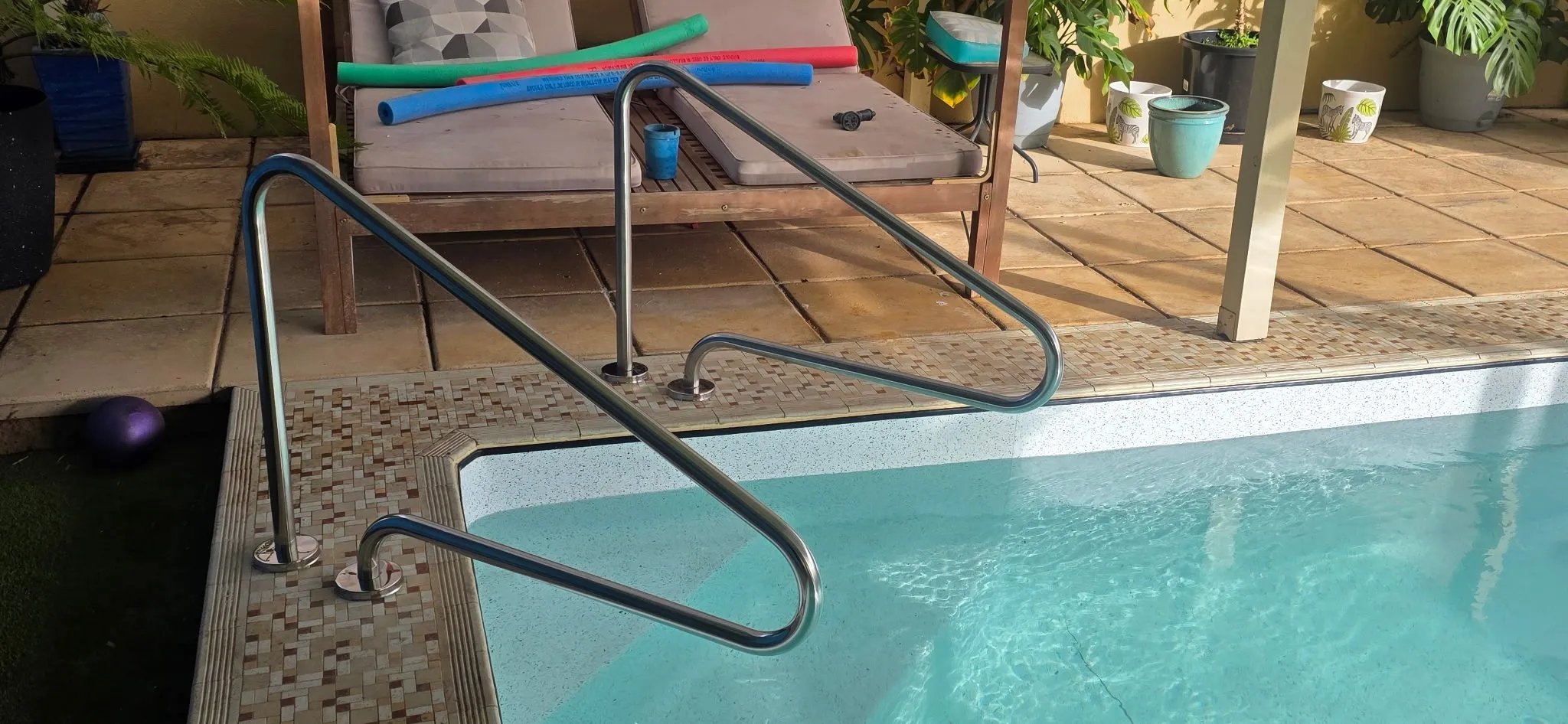 Daytime detail of SR Smith handrails mounted on mosaic tile coping with daybed and tropical planting beyond
